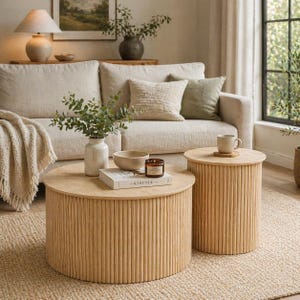 May include: Two light wood coffee tables with a ribbed design. The round tables are different sizes and sit on a woven rug. A sofa with neutral-colored pillows and a vase of greenery are in the background.