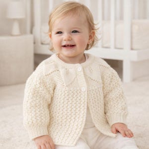 May include: Cream-colored knitted baby cardigan with a textured stitch pattern. The cardigan features a round neckline, button closure, and decorative bow details on the shoulders. The baby is wearing the cardigan.
