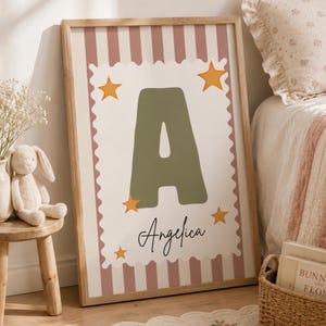 Puede incluir: Un estampado enmarcado con una gran letra "A" verde oliva y el nombre "Angelica" en cursiva. El fondo tiene rayas rosas y blancas, con acentos de estrellas naranjas. El estampado está en un marco de madera clara.