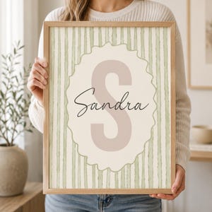 Może przedstawiać: Oprawiony wydruk z imieniem "Sandra" w eleganckim skrypcie nad dużą literą "S". Projekt zawiera falisty obramowanie i pasiasty tło w miękkich odcieniach zieleni i kremu. Rama wykonana jest z jasnego drewna.