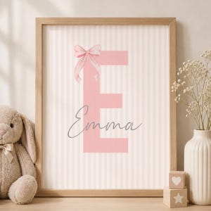 Könnte beinhalten: Ein gerahmter Druck mit einem rosa Buchstaben "E" mit Schleife, dem Namen "Emma" in Schreibschrift und einem gestreiften Hintergrund. Der Rahmen ist hellbraun. Ein Stoffhase, eine Vase mit Blumen und Holzblöcke sind ebenfalls zu sehen.