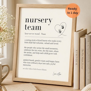 以下が含まれることがあります： 「nursery team」という黒いフォントの言葉が書かれた額入りのプリント。プリントには定義、手形のあるハート、そして「Ready In 1 day」というテキストが含まれています。フレームはライトブラウンです。