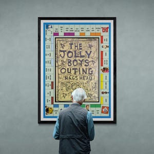 Op de afbeelding: Een ingelijst kunstwerk met een Monopoly-bordspelontwerp met de tekst "THE JOLLY BOYS OUTING NAGS HEAD PECKHAM" in het midden. Het kunstwerk staat in een zwart frame en wordt tentoongesteld op een grijze muur. Een persoon staat voor het kunstwerk.