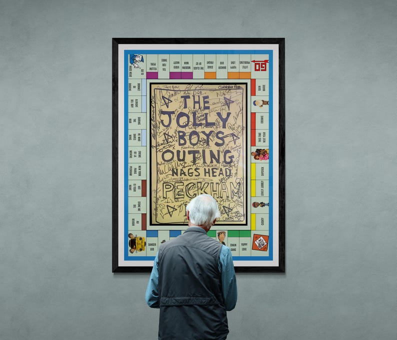Op de afbeelding: Een ingelijst kunstwerk met een Monopoly-bordspelontwerp met de tekst "THE JOLLY BOYS OUTING NAGS HEAD PECKHAM" in het midden. Het kunstwerk staat in een zwart frame en wordt tentoongesteld op een grijze muur. Een persoon staat voor het kunstwerk.