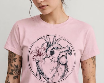 Może przedstawiać: Jasnoróżowy t-shirt z krótkim rękawem z okrągłym nadrukiem graficznym na klatce piersiowej. Wzór przedstawia anatomiczne serce splecione z kwiatem lilii i drutem kolczastym, wykonane ciemnymi liniami tuszu wewnątrz cienkiej okrągłej ramki.