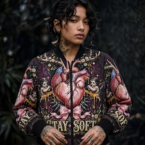 Könnte beinhalten: Eine schwarze Bomberjacke mit einem detaillierten Grafikdruck eines zentralen anatomischen Herzens, Putten, Geweihen und Gänseblümchen. Das Design umfasst eine rosa Schleife und den Schriftzug "STAY SOFT" in goldenen Buchstaben auf der Vorderseite.