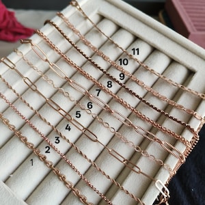 Puede incluir: Una variedad de once collares de oro rosa exhibidos sobre una superficie crema. Los collares presentan varios estilos de cadena, incluyendo diseños de cable, cuerda y clip. Cada collar está numerado para una fácil identificación, mostrando la variedad de opciones disponibles.