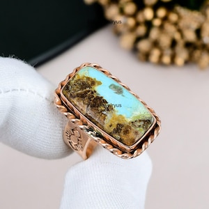 May include: A copper-colored ring with a rectangular turquoise stone. The stone has brown and blue hues. The ring has a twisted rope design around the stone and a decorative design on the band.