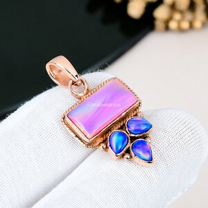 May include: Rose gold pendant featuring a rectangular pink opal and three blue opal teardrop accents. The rectangular opal has a rope-like border. The pendant is on a white surface. Text on the pendant reads "Elarajewelryus".