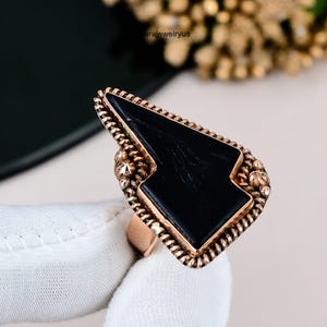May include: A rose gold ring featuring a large, black, geometric stone. The stone is set in a decorative frame with a beaded edge. The ring is held against a white background. The text "arajewelryus" is visible.