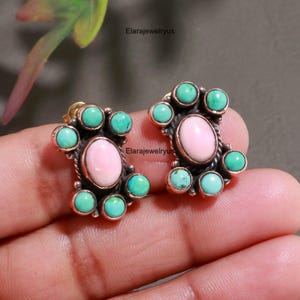 May include: A pair of stud earrings featuring a central oval pink stone surrounded by six small round turquoise stones set in a dark metal frame. The text Elarajewelryus is visible on the image. The design includes a twisted metal detail.