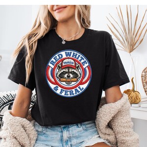 Może przedstawiać: Czarny t-shirt z okrągłą grafiką przedstawiającą szopa w kapeluszu trzymającego hot doga. Grafika zawiera napisy "Red, White & Feral" i "'merica". Projekt jest w kolorach czerwonym, białym i niebieskim.