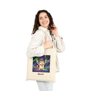 May include: A beige tote bag featuring a colorful illustration of a cartoon cat wearing a hat and holding glowing sticks. The cat is in front of a neon green and purple background. The word "Ravin'" is printed at the bottom of the image.