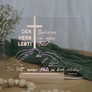 Könnte beinhalten: Eine klare Acryl-LED-Leuchte mit geätztem Text und einem Kreuzdesign. Der Text enthält deutsche Sätze wie "DER HERR LEBT!". Die Leuchte steht auf einem Holzsockel, mit einer Bergkontur und dem Bibelvers Psalm 18,47.