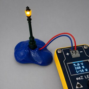 May include: A miniature street lamp with a glowing yellow light, connected by red and blue wires to a yellow and black electronic device. The device displays voltage, resistance, and wattage. The lamp is mounted on a blue base.