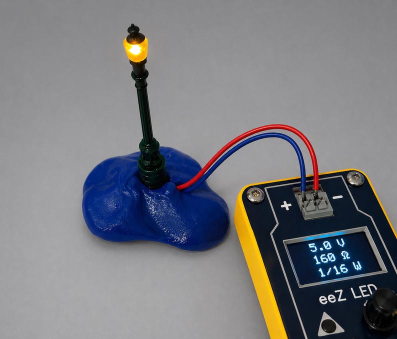 May include: A miniature street lamp with a glowing yellow light, connected by red and blue wires to a yellow and black electronic device. The device displays voltage, resistance, and wattage. The lamp is mounted on a blue base.