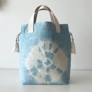 May include: A light blue tote bag with a white floral pattern. The bag has cream-colored handles and tassels on the sides. The bag is made of a soft, textured fabric.