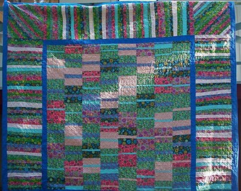 May include: A rectangular quilt featuring a central panel of alternating vertical strips in shades of pink, green, blue, and purple with floral patterns. The center is framed by a solid blue border and an outer border of horizontal stripes.