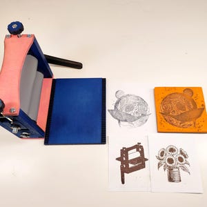 Könnte beinhalten: Eine kleine Tischdruckpresse in Blau, Rosa und Grau mit einem schwarzen Griff. Es werden mehrere Drucke gezeigt, darunter ein Blumendesign, ein Himmelsbild und ein Bär. Die Drucke sind in Braun- und Schwarztönen gehalten.