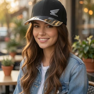 May include: A black baseball cap featuring a silver Honda logo on the front panel and a metallic silver brim. The cap has a structured crown and a curved visor, displaying the brand name Honda in white text beneath the silver wing emblem.
