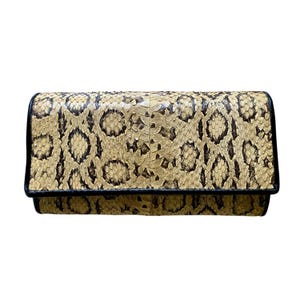 May include: A rectangular clutch wallet featuring a textured snakeskin pattern in shades of beige, tan, and dark brown. The wallet has a fold-over flap design with a solid black leather trim along the edges, providing a defined border.