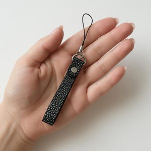 Könnte beinhalten: Eine schwarze und graue Handschlaufe mit strukturiertem Muster und einem silberfarbenen Metallring und Knopf. Die Schlaufe hat eine schwarze Kordelschlaufe zur Befestigung an einem Gerät.