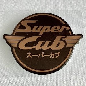 Op de afbeelding: Een ronde sticker met een donkerbruine achtergrond, een lichtbruine rand en gestileerde vleugels. De tekst Super Cub is in het lichtbruin gedrukt, met daaronder de Japanse katakana-tekens voor Super Cub in dezelfde lichtbruine kleur.