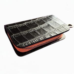以下が含まれることがあります： クロコダイル柄の黒い財布。財布にはコーラル色のジッパーと、側面に付いたシルバー色の金属リングが付いています。財布は長方形で、革製のように見えます。