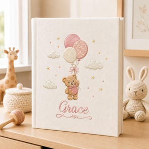 Può includere: Un libro dei ricordi rivestito in tessuto bianco con un ricamo raffigurante un orsetto marrone che tiene tre palloncini nei toni del rosa e del crema. Il ricamo include nuvole, stelle e il nome Grace in corsivo rosa sotto l'orsetto.