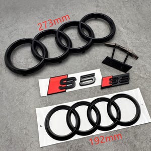 May include: Black car emblem set, including the Audi rings measuring 273mm and 192mm, and S5 badges with red accents. The set also includes a mounting bracket. The items are displayed on a gray surface.