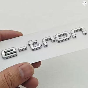 Könnte beinhalten: Ein verchromtes "e-tron"-Emblem, wahrscheinlich für ein Fahrzeug, wird auf weißem Hintergrund gezeigt. Die Buchstaben sind metallisch und haben eine dreidimensionale Optik. Das Emblem wird von einer Hand gehalten.