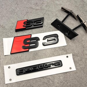 Op de afbeelding: Zwarte S3- en Quattro-auto-emblemen met rode accenten. De S3-emblemen hebben een 3D-ontwerp. Ook inbegrepen zijn een paar zwarte manchetknopen met zilveren accenten. De items liggen op een wit oppervlak.