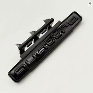 May include: A black Quattro emblem with a glossy finish. The emblem has a rectangular shape with the word "Quattro" embossed in a bold, sans-serif font. The emblem has a mounting bracket on the back.