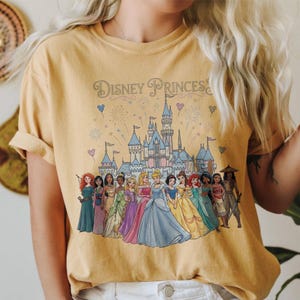 K&ouml;nnte beinhalten: Senfgelbes T-Shirt mit dem Text "Disney Princesses" &uuml;ber einer Grafik eines Schlosses und mehrerer Disney-Prinzessinnen. Die Prinzessinnen tragen farbenfrohe Kleider. Feuerwerk und Herzen sind ebenfalls im Design enthalten.