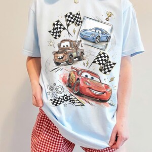 May include: Light blue t-shirt featuring cartoon cars, including a red race car, a brown tow truck, and a blue sports car. The design includes checkered flags and a trophy. The shirt is paired with red and white checkered shorts.
