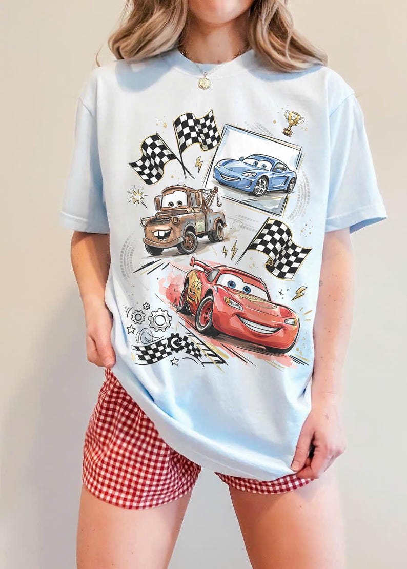 May include: Light blue t-shirt featuring cartoon cars, including a red race car, a brown tow truck, and a blue sports car. The design includes checkered flags and a trophy. The shirt is paired with red and white checkered shorts.