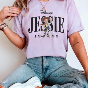 May include: A light purple t-shirt featuring the Disney logo and the name "JESSIE" in large black letters, with the number "1999" below. The shirt has a graphic of the character Jessie from Toy Story. The person is wearing blue jeans and white sneakers.
