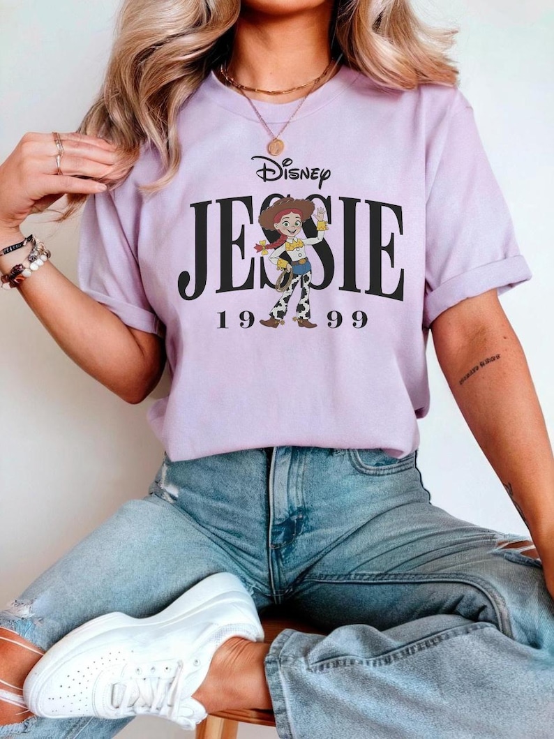 May include: A light purple t-shirt featuring the Disney logo and the name "JESSIE" in large black letters, with the number "1999" below. The shirt has a graphic of the character Jessie from Toy Story. The person is wearing blue jeans and white sneakers.