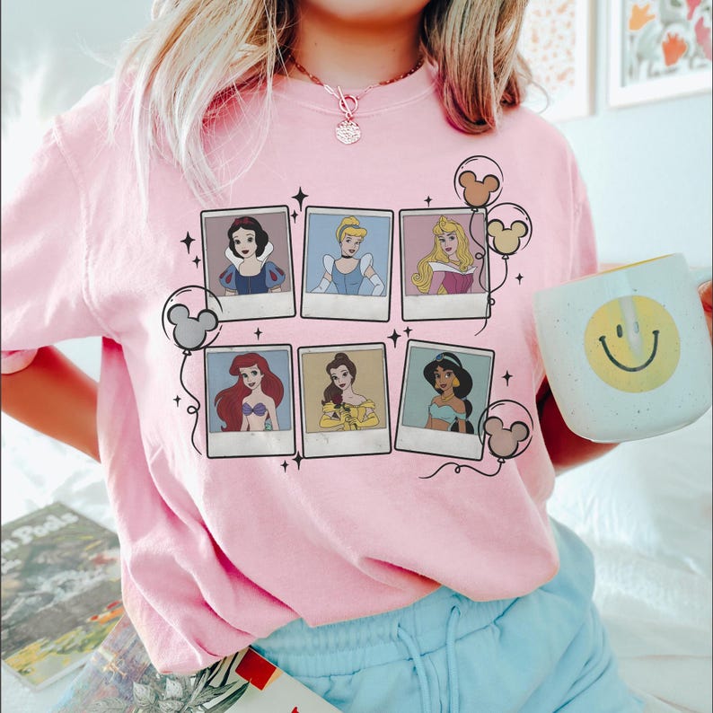 May include: Pink t-shirt featuring six Disney princess portraits in polaroid frames. Mickey Mouse balloon graphics and a smiley face mug are also visible. The shirt is a light pink color.
