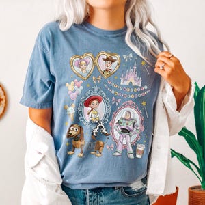 May include: A pale blue t-shirt with a colourful design featuring characters from the Toy Story film series. The design includes images of Woody, Buzz Lightyear, Jessie, and other characters, with the phrase "To Infinity and Beyond."