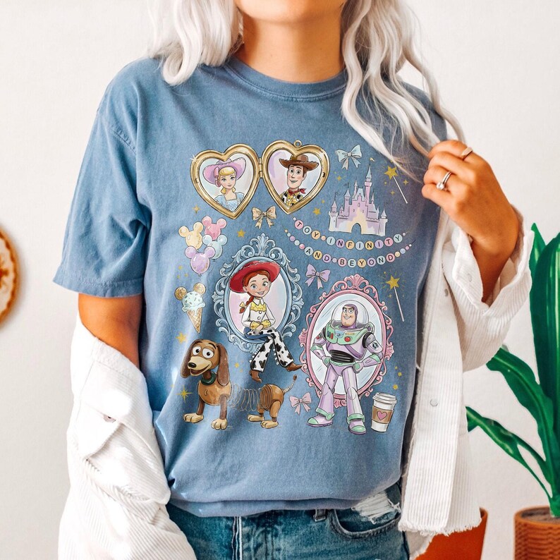 May include: A pale blue t-shirt with a colourful design featuring characters from the Toy Story film series. The design includes images of Woody, Buzz Lightyear, Jessie, and other characters, with the phrase "To Infinity and Beyond."