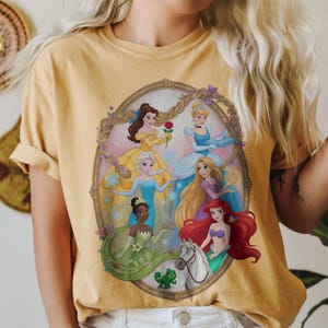 May include: Mustard yellow t-shirt with a framed illustration of Disney princesses. The image shows princesses in their signature gowns, including Belle with a rose and Ariel with red hair. The tee has short sleeves and a crew neck.