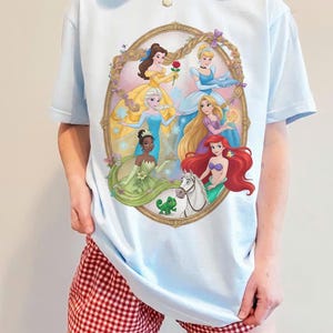 May include: Pale blue t-shirt with a framed illustration of Disney princesses. The artwork includes Belle, Elsa, Cinderella, Rapunzel, Ariel, and Tiana. The shirt is worn with red and white checked shorts.