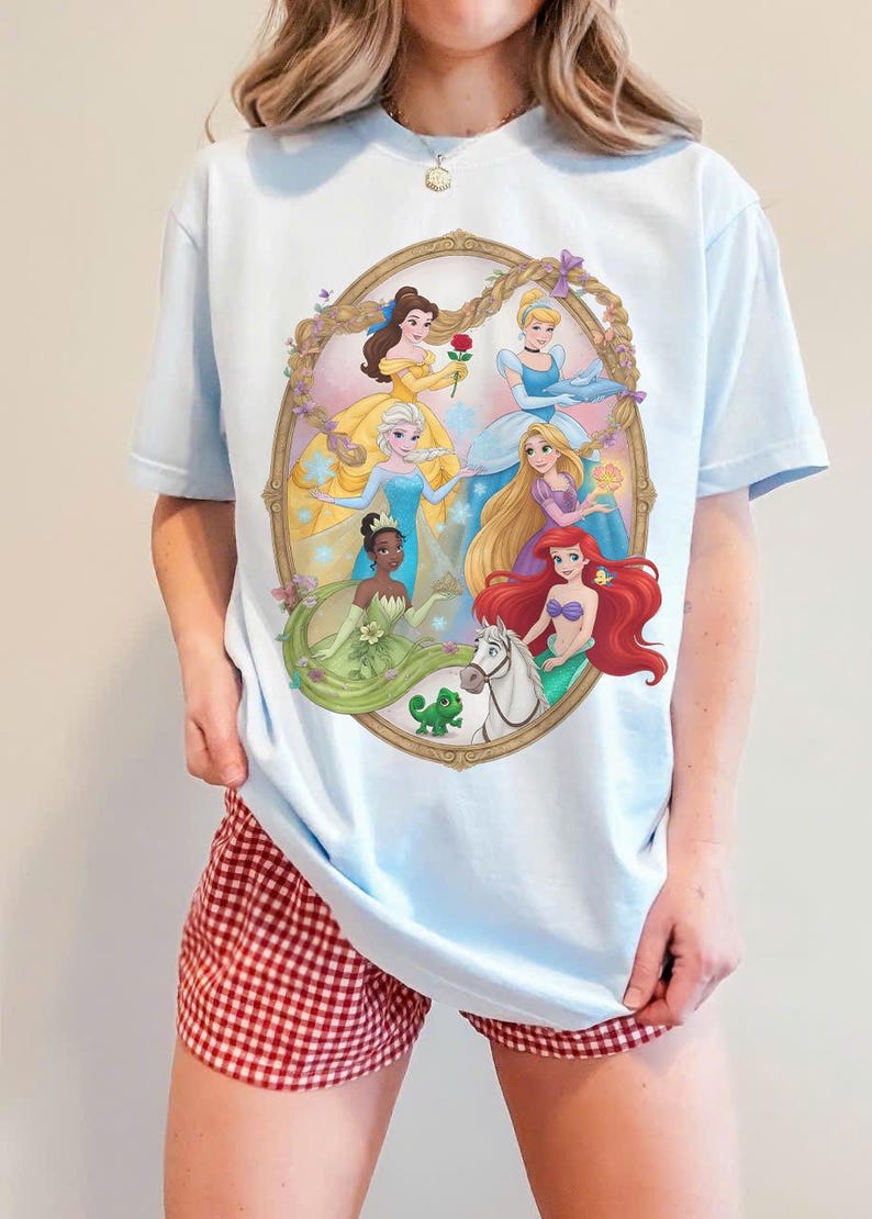 May include: Pale blue t-shirt with a framed illustration of Disney princesses. The artwork includes Belle, Elsa, Cinderella, Rapunzel, Ariel, and Tiana. The shirt is worn with red and white checked shorts.