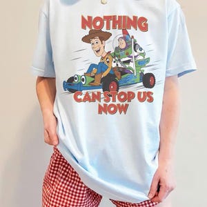 May include: Light blue t-shirt featuring a graphic of Woody, Buzz Lightyear, and a toy car with the text "NOTHING CAN STOP US NOW" in red. The shirt is a light blue color.