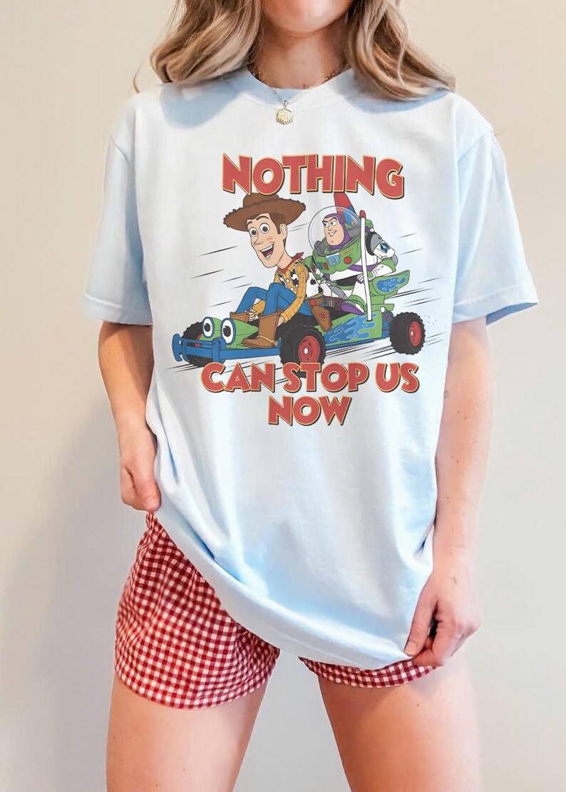 May include: Light blue t-shirt featuring a graphic of Woody, Buzz Lightyear, and a toy car with the text "NOTHING CAN STOP US NOW" in red. The shirt is a light blue color.