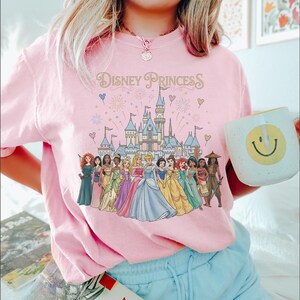 K&ouml;nnte beinhalten: Hellrosa T-Shirt mit dem Schriftzug "Disney Princess" und einem Schloss. Das T-Shirt zeigt Illustrationen von Disney-Prinzessinnen. Eine wei&szlig;e Tasse mit einem gelben Smiley wird in der Hand gehalten.