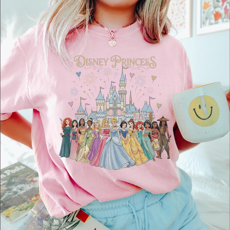 K&ouml;nnte beinhalten: Hellrosa T-Shirt mit dem Schriftzug "Disney Princess" und einem Schloss. Das T-Shirt zeigt Illustrationen von Disney-Prinzessinnen. Eine wei&szlig;e Tasse mit einem gelben Smiley wird in der Hand gehalten.