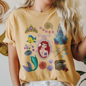Puede incluir: Camiseta de color mostaza con un gr&aacute;fico colorido de Ariel de La Sirenita. El dise&ntilde;o incluye una corona, Flounder, una concha, un castillo, una cola de sirena y un cofre del tesoro. La camiseta tiene mangas cortas y cuello redondo.