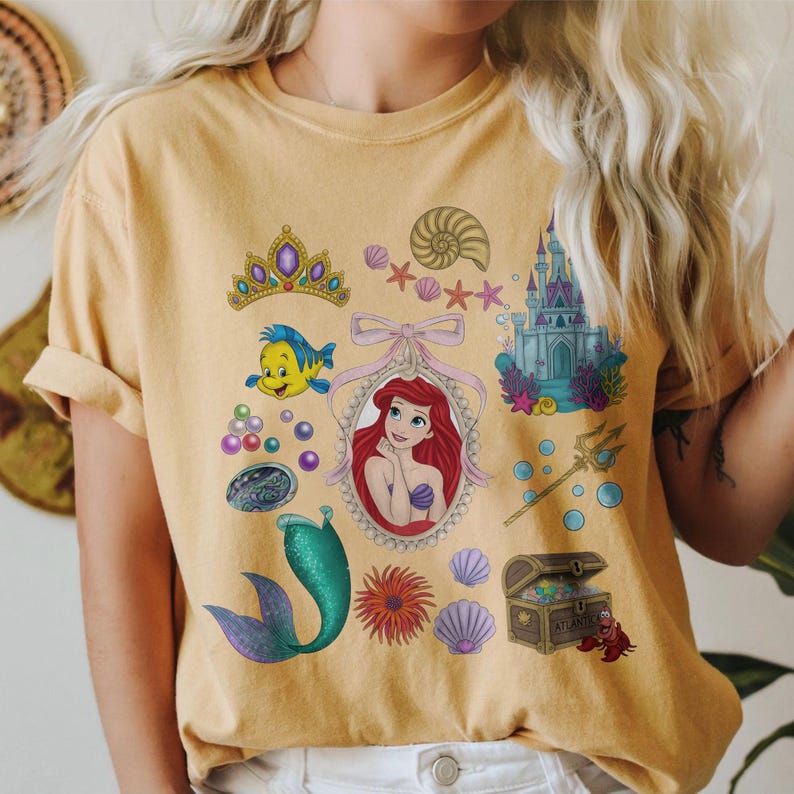 Puede incluir: Camiseta de color mostaza con un gr&aacute;fico colorido de Ariel de La Sirenita. El dise&ntilde;o incluye una corona, Flounder, una concha, un castillo, una cola de sirena y un cofre del tesoro. La camiseta tiene mangas cortas y cuello redondo.